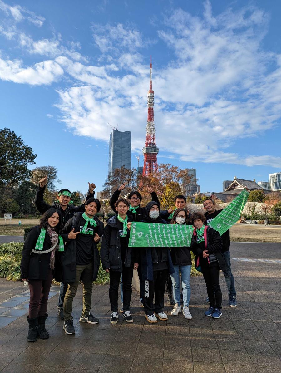 10 New Relic employees wearing New Relic Volunteer T-shirts stand in a Tokyon park smiling and holding New Relic banners.